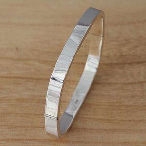 May include: A polished silver bangle bracelet with a flat, rectangular design. The bracelet has a smooth surface and reflects light. The bracelet is displayed on a light brown wooden surface. The bracelet appears to be made of metal.