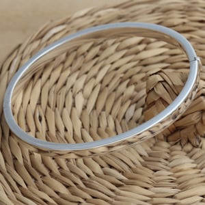 May include: A silver bangle bracelet with a simple, elegant design. The bracelet is round and has a smooth, polished surface. The clasp is visible on the side. The bracelet is displayed on a woven, light brown surface.