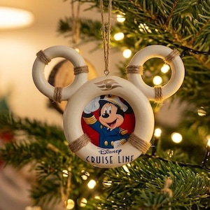 May include: A cream-colored life preserver ornament shaped like Mickey Mouse ears, featuring a central image of Mickey Mouse in a captain's uniform. The text reads Disney Cruise Line. It is suspended by a thin brown twine string.
