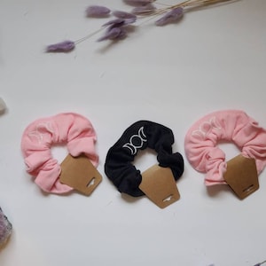 May include: Three hair scrunchies with a moon phase design. One is pink, one is black, and one is light pink. Each scrunchie has a brown paper tag attached.