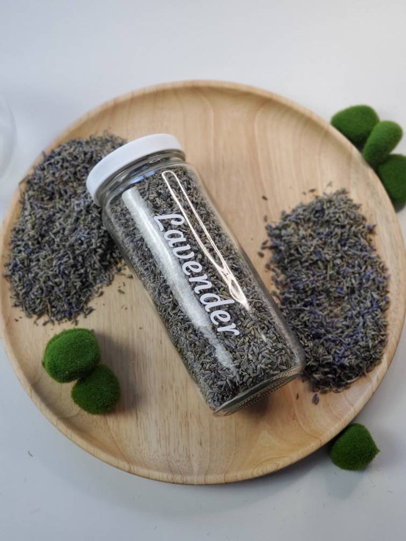 Puede incluir: Un frasco de vidrio lleno de flores de lavanda secas, etiquetado como "Lavender", sobre una bandeja de madera con bolas de musgo verde.