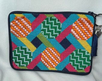A finished needlepoint purse/cosmetic bag with a colorful patchwork design. It has a navy blue tassel, an antique heart and a “word” charm.