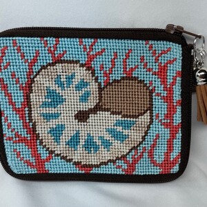 May include: A small blue and white needlepoint zippered pouch with a brown tassel. The pouch features a nautilus shell design with coral branches in the background.