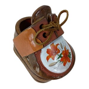 May include: A ceramic baby shoe with a brown and tan color scheme. The shoe features a painted design of orange lilies on a white background. The shoe has a leather strap and laces.