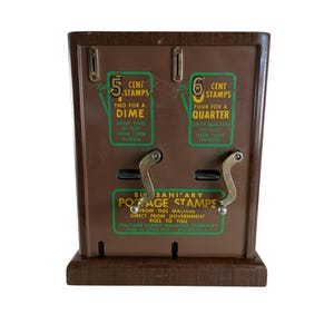 May include: A vintage brown postage stamp vending machine with two dispensing slots. The machine has instructions for 5 and 6 cent stamps, with handles and text that reads "Buy Sanitary Postage Stamps".