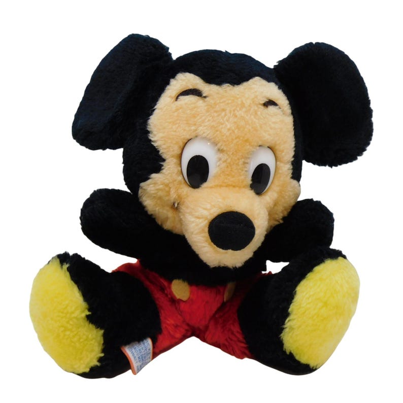 Może przedstawiać: Pluszowa zabawka Mickey Mouse siedząca ze skrzyżowanymi nogami. Zabawka ma na sobie czerwone spodnie z ż&oacute;łtym guzikiem i ma czarne futro z ż&oacute;łtymi butami.