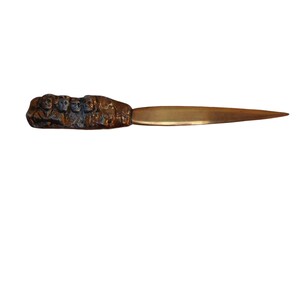 May include: A brass letter opener with a detailed design of several figures on the handle. The blade is a shiny gold color.
