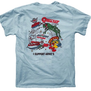 May include: Light blue t-shirt with a graphic print of various retro arcade game characters, including Pac-Man, Centipede, and Asteroids. The text "I Support Arnie's" is printed below the graphic.
