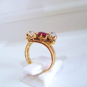 May include: A gold ring featuring a three-stone setting with a central deep red gemstone flanked by two clear, faceted white gemstones. The stones are held in place by decorative metal prongs, and the band has a smooth, polished gold finish.