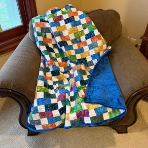 May include: A colorful patchwork quilt draped over a brown armchair. The quilt features a vibrant pattern of squares in various colors, including blue, orange, yellow, and purple. The underside is a solid blue, plush fabric.