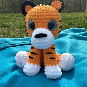 May include: A crocheted orange and black tiger plush toy with white paws and blue eyes. The toy is sitting on a blue blanket.