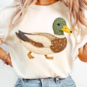 Könnte beinhalten: Cremefarbenes T-Shirt mit einem skurrilen Enten-Design. Die Ente hat einen grün karierten Kopf, einen gelben Schnabel und einen Körper, der aus verschiedenen Stoffmustern besteht, darunter florale und strukturierte Elemente. Die Ente geht.