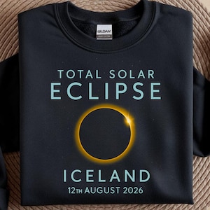 May include: Black sweatshirt featuring the text "TOTAL SOLAR ECLIPSE ICELAND 12th AUGUST 2026" in light blue. A yellow and black solar eclipse graphic is centered on the front. The sweatshirt is displayed on a woven surface.
