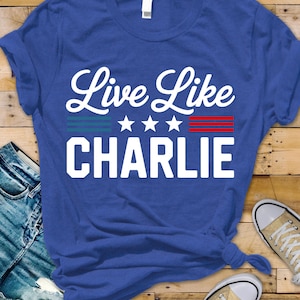 May include: A royal blue t-shirt with the text "Live Like CHARLIE" in white script. The design includes three white stars and blue and red stripes. The shirt is displayed on a wooden surface with jeans and sneakers.
