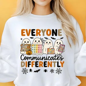 May include: White sweatshirt with the text "EVERYONE COMMUNICATES DIFFERENTLY" in brown and orange. Four cartoon ghosts hold up a pumpkin, an alphabet chart, and a phone. Halloween-themed apparel.