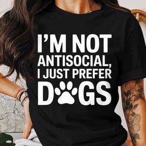 Peut inclure: T-shirt noir avec le texte blanc "I'M NOT ANTISOCIAL, I JUST PREFER DOGS" et un motif de patte. Le t-shirt est à col rond et à manches courtes. Le thème général est celui des amoureux des chiens.