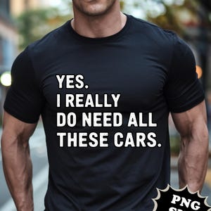 May include: Black t-shirt with the text "YES. I REALLY DO NEED ALL THESE CARS." in white block letters. The shirt is short-sleeved and appears to be made of a soft material. The background is blurred.
