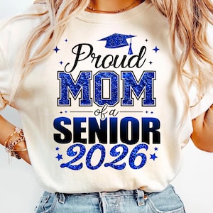 May include: Cream-colored t-shirt with the text "Proud MOM of a SENIOR 2026" in blue glitter lettering. A graduation cap graphic is above the text. The shirt is a casual style.
