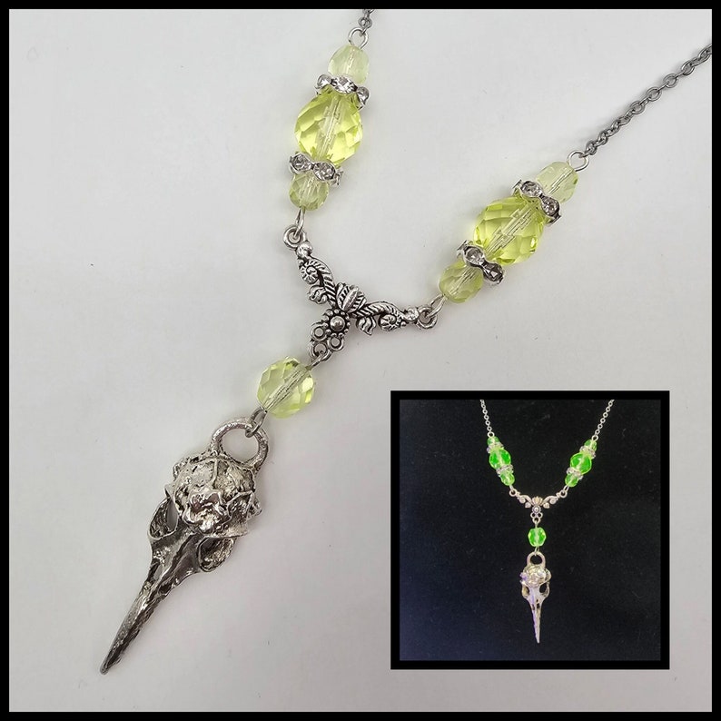 May include: A silver necklace with a bird skull pendant and green glass beads. The necklace is on a white background. The pendant is detailed and has a textured surface. The beads are faceted and have a light green color.