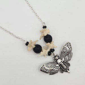 May include: A silver chain necklace with a silver moth pendant featuring a skull design. The necklace also has black lava beads and white bone beads.
