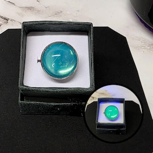 May include: A round ring with a large, iridescent teal stone set in a silver-colored metal band. The ring is displayed in a small, dark green velvet box with a white interior, set on a black surface. A smaller, illuminated version of the ring is also shown.