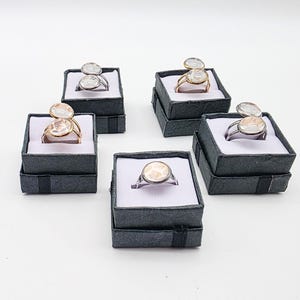 May include: Five rings presented in individual black gift boxes with white interiors. The rings feature clear, oval-shaped stones. Some rings have two stones, while others have one. The ring bands are gold or silver.