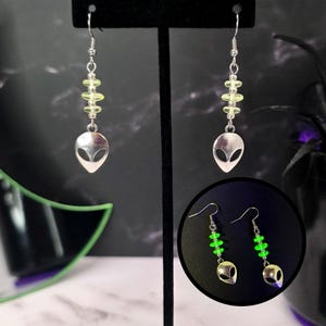 May include: Silver alien head dangle earrings with a series of light green beads. The earrings have a hook closure. The image also shows a close-up of the earrings with a glowing green effect.