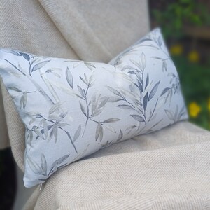 May include: A light blue throw pillow with a gray and white leafy pattern. The pillow is sitting on a beige throw blanket.