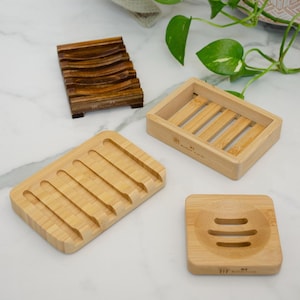 May include: Three wooden soap dishes made of bamboo. The dishes are all different shapes and sizes. The first dish is a long, rectangular dish with five ridges. The second dish is a square dish with a rounded edge and three ridges. The third dish is a rectangular dish with six ridges. All three dishes have the words "Mother Earth" printed on the bottom.