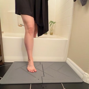 May include: A rectangular, two-piece gray bath mat featuring a geometric, embossed line pattern. The mat is composed of two interlocking sections that lie flat on the floor, providing a stable surface for stepping out of a bathtub or shower area.