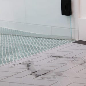 May include: A close-up of a grey and white patterned bath mat with a dark grey footprint on it. The mat is placed on top of a light green tiled floor.