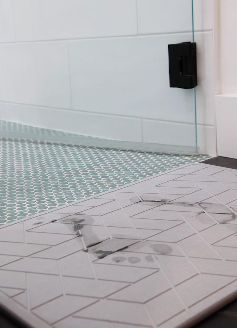 May include: A close-up of a grey and white patterned bath mat with a dark grey footprint on it. The mat is placed on top of a light green tiled floor.