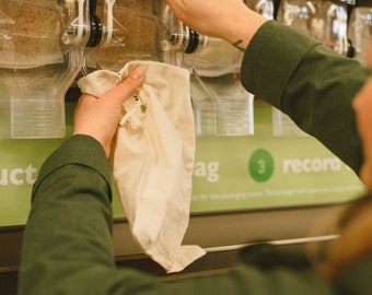 May include: A person is filling a white reusable bag with brown rice from a bulk bin dispenser. The bin is labeled "Organic Short Grain Brown Rice" and has a price tag of $2.89 per pound. The dispenser is part of a row of bulk bins with various grains and other products.