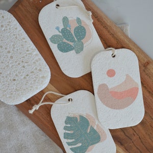 May include: Three rectangular, white, textured ceramic tags with string loops. Each tag features a different botanical illustration: a cactus, a mountain landscape, and a monstera leaf. The illustrations are in shades of green, pink, and brown.