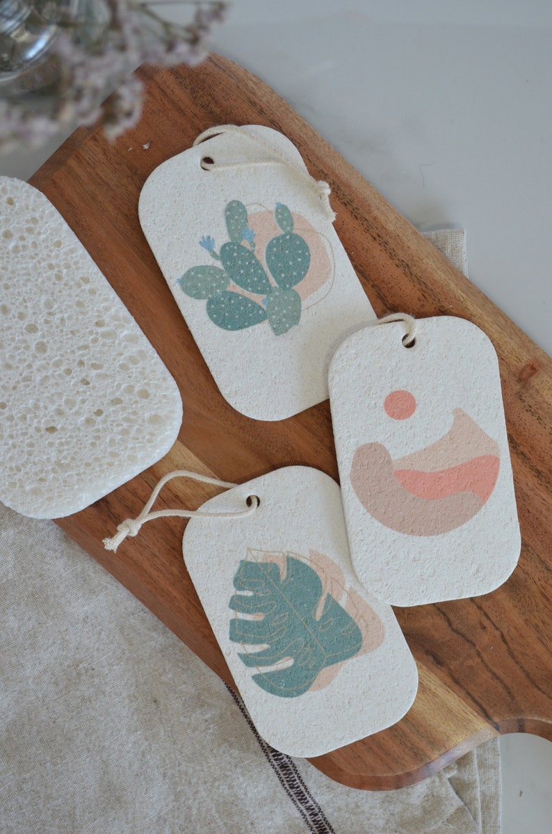 May include: Three rectangular, white, textured ceramic tags with string loops. Each tag features a different botanical illustration: a cactus, a mountain landscape, and a monstera leaf. The illustrations are in shades of green, pink, and brown.