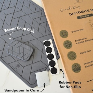 May include: A dark grey diatomite mat with a geometric pattern. The mat has a bonus soap dish and rubber pads for non-slip. The mat is labelled "Quick-Dry Diatomite Mat" and "Dark Grey".