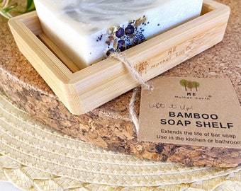 May include: A bamboo soap shelf with a bar of soap on top. The soap is white with gray swirls and black seeds. The shelf is labeled "Lift it Up! Bamboo Soap Shelf" and "ME Mother Earth".