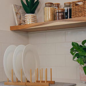 May include: A white ceramic dish rack with wooden dowels, holding three white plates. The rack is on a grey countertop. A wooden shelf with jars of grains and spices is above the rack. A potted succulent plant is on the shelf. The text "Living without plastic" is visible on a book on the shelf.