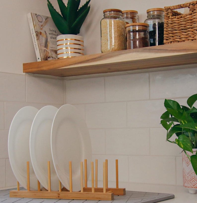 May include: A white ceramic dish rack with wooden dowels, holding three white plates. The rack is on a grey countertop. A wooden shelf with jars of grains and spices is above the rack. A potted succulent plant is on the shelf. The text "Living without plastic" is visible on a book on the shelf.