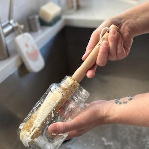 May include: A long-handled bottle brush with light-colored wooden handle and natural tan bristles inserted into a clear glass jar. The brush head is covered in white soap suds, and a small white cord loop is attached to the end of the wooden handle.