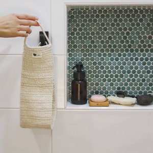 May include: A white bath pouf hanging on a hook in a bathroom. The pouf is made of a textured material and has a green leaf logo on it. The bathroom has white tiles and a green mosaic tile shower niche with a bottle of soap, a bar of soap, and a loofah.