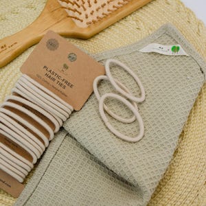 Op de afbeelding: Een houten haarborstel, een verpakking van twintig beige plasticvrije haarelastiekjes met het label 100% Cotton and Natural Rubber, en een saliegroene doek met wafelstructuur. Het handvat van de borstel bevat de tekst ME Mother Earth.