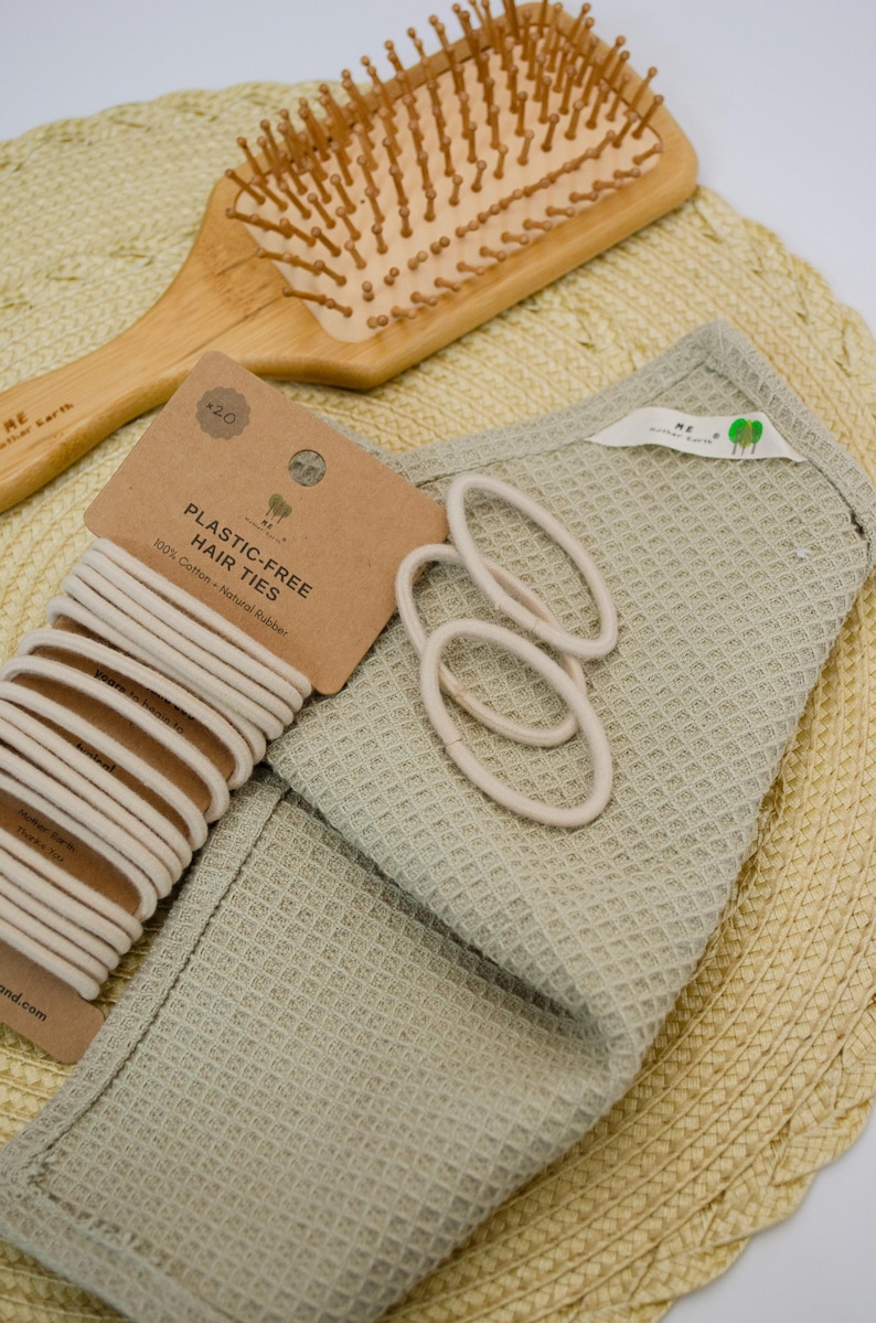 Op de afbeelding: Een houten haarborstel, een verpakking van twintig beige plasticvrije haarelastiekjes met het label 100% Cotton and Natural Rubber, en een saliegroene doek met wafelstructuur. Het handvat van de borstel bevat de tekst ME Mother Earth.