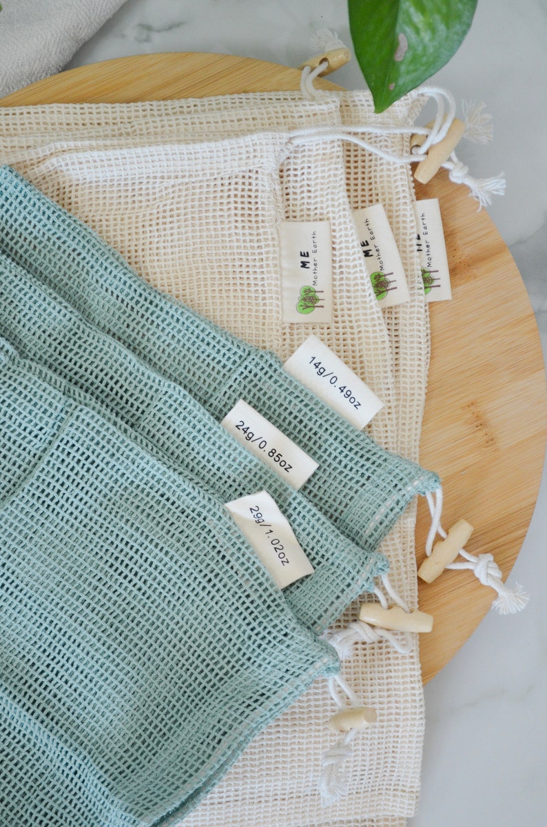 May include: Three reusable mesh produce bags in different sizes. The bags are made of a fine mesh material and have a drawstring closure. The bags are labeled with their weight in ounces and grams. The bags are a light blue color and a natural white color.