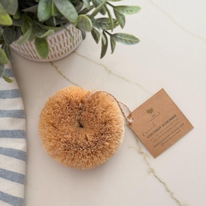 May include: A round, tan coconut scrubbie with a tag that reads "Coconut Scrubbie" and "memotherearthbrand.com". The scrubbie is made with coconut fiber bristles. A small potted plant and a blue and white striped towel are in the background.
