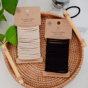 Op de afbeelding: Twee rechthoekige kartonnen displaykaarten met plasticvrije haarelastiekjes, &eacute;&eacute;n met beige elastiekjes en &eacute;&eacute;n met zwarte. Elke kaart is gelabeld met PLASTIC-FREE HAIR TIES 100% Cotton + Natural Rubber en bevat de website memotherearthbrand.com.