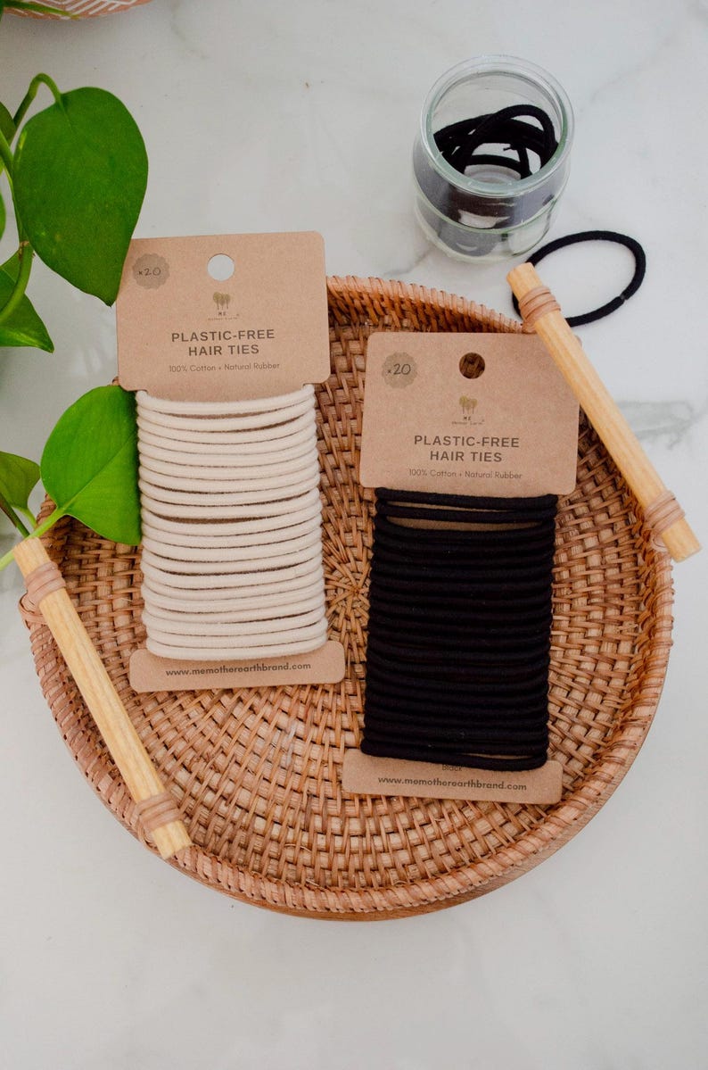 Op de afbeelding: Twee rechthoekige kartonnen displaykaarten met plasticvrije haarelastiekjes, &eacute;&eacute;n met beige elastiekjes en &eacute;&eacute;n met zwarte. Elke kaart is gelabeld met PLASTIC-FREE HAIR TIES 100% Cotton + Natural Rubber en bevat de website memotherearthbrand.com.