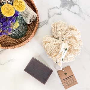 May include: A bar of dark purple soap and a beige sisal shower pouf with a tag that reads "Reusable Sisal Shower Pouf". A woven basket with flowers and a rolled towel is in the background. The items are on a white marble surface.