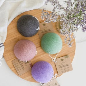 May include: Four round konjac facial cleansing sponges in charcoal grey, dusty pink, light green, and lavender purple. Each sponge features a white hanging string and a small brown cardboard tag labeled with the product name and brand logo.
