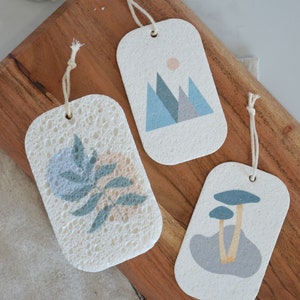 May include: Three white, rectangular, textured, hanging ornaments with different designs. One ornament features a blue and green leaf, another features a blue and gray mountain range, and the last features a gray mushroom with a yellow stem.